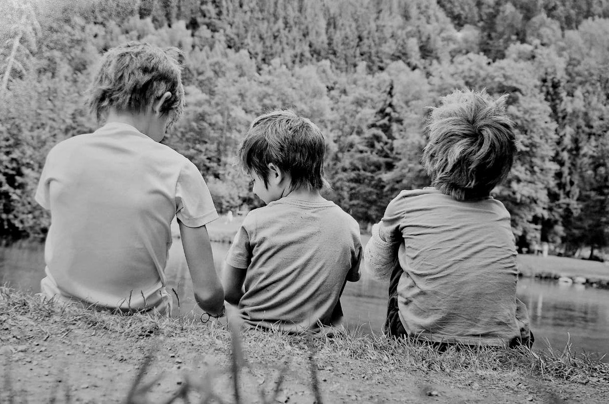 Trois enfants assis dans l’herbe, de dos, face à l’eau et à la forêt
